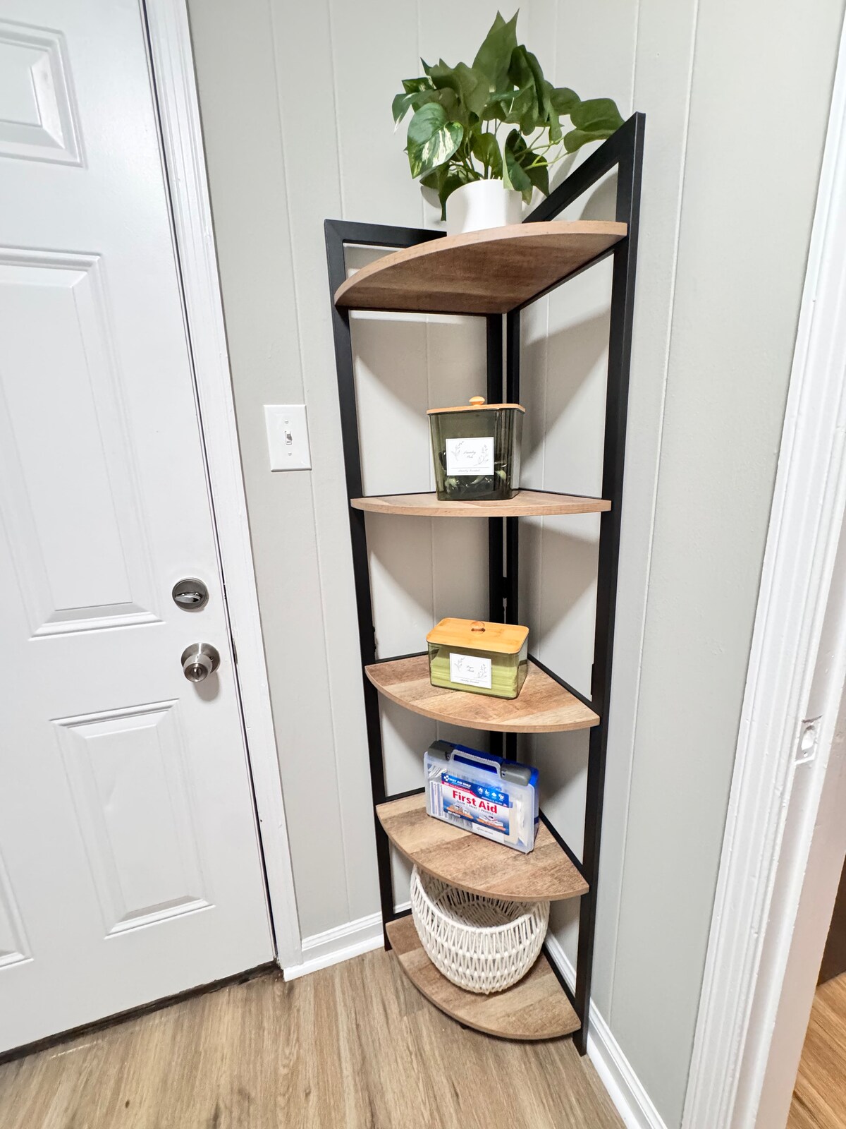 A corner shelving unit features five wooden shelves with a natural finish, displaying various storage containers and a potted plant at the top. The unit stands against a light gray wall, next to a white door, contributing to a neat and organized entryway.