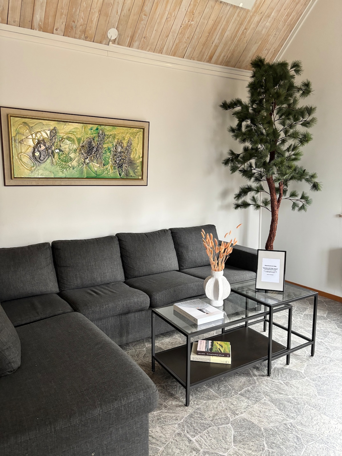 A comfortable seating area is featured, highlighted by a dark gray sectional sofa. A glass coffee table holds decorative items, including a vase with dried flowers. An abstract artwork adorns the wall, while a faux tree adds a touch of greenery to the space.