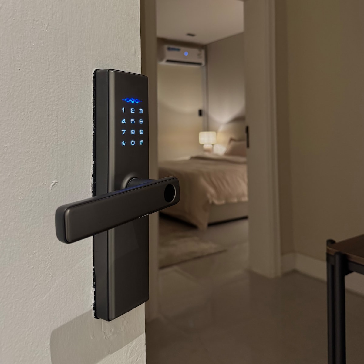 A modern electronic keypad lock is visible on a white wall, providing secure access to the interior. In the background, a softly lit bedroom is partially visible, featuring a plush bed and bedside lamps that create a calm ambiance.