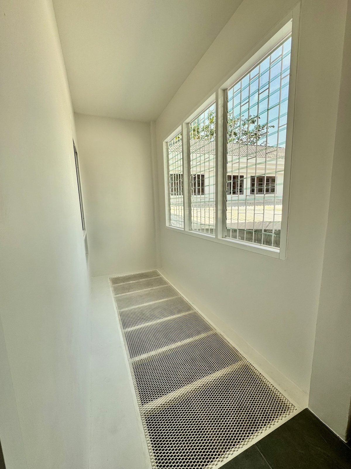 A bright hallway features large windows on one side, allowing natural light to fill the space. The white walls create an airy feel, while a grid-patterned floor adds texture. Ventilation grates line the floor, contributing to a contemporary design.