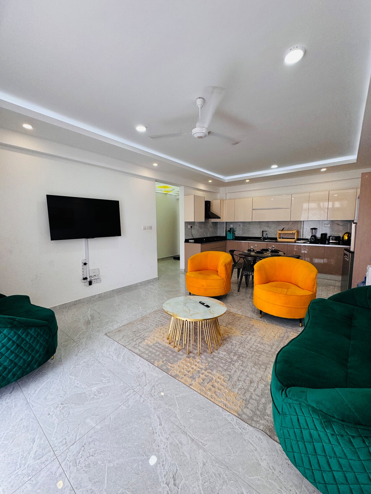 A modern living area features two vibrant yellow armchairs and a plush green sofa, inviting relaxation. A round marbled coffee table sits at the center, complemented by a subtle area rug. A wall-mounted television is positioned above a sleek kitchen with contemporary cabinets visible in the background.
