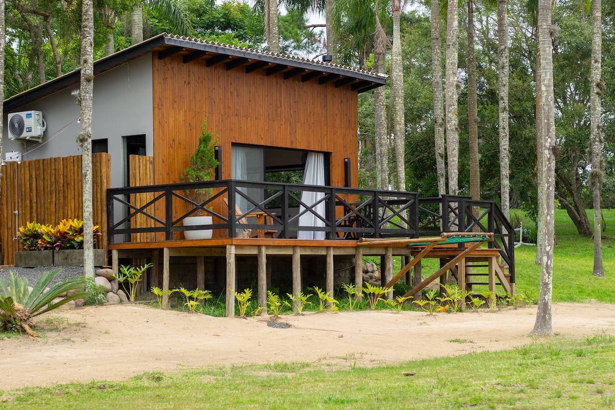 A wooden cabin features a large deck with distinct black railings, surrounded by lush greenery and trees. A few comfortable chairs are placed on the deck, inviting relaxation. The cabin's façade showcases a mix of natural wood and modern design, harmonizing with the tranquil outdoor setting.