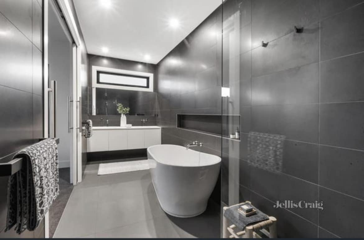 A modern bathroom is highlighted by sleek dark tiles and minimalist design. A freestanding bathtub is positioned near a glass shower, while a white vanity provides ample storage. Soft lighting enhances the atmosphere, creating a serene space for relaxation.