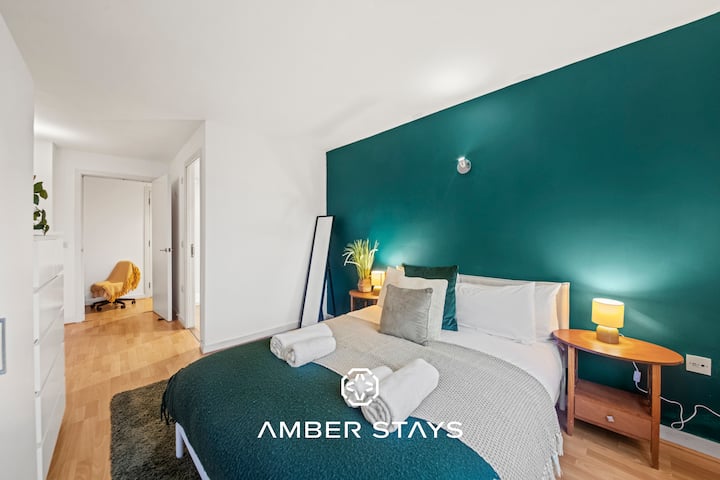 Ova svijetla, moderna spavaća soba ima bijele zidove i namještaj, uključujući veliki ormar i komodu, naglašene tirkiznom i sivom posteljinom. Veliki prozor ispunjava prostor prirodnim svjetlom, pružajući jasan pogled na gradske zgrade.