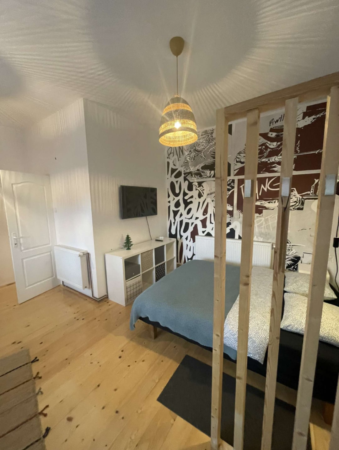 A well-lit bedroom features a wooden divider offering privacy. A large bed is dressed in a soft blue blanket with silver pillows. A television is mounted on the wall across from the bed. Artistic wall decor and a pendant light add character to the space.