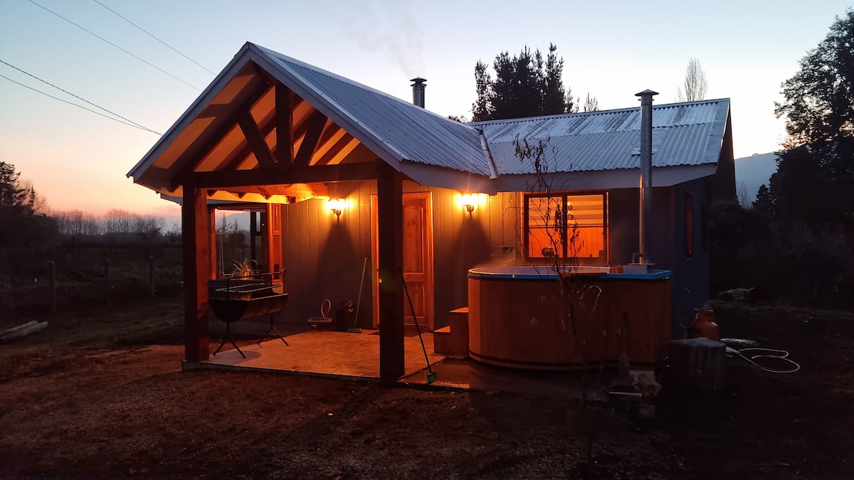 The exterior of a charming house is illuminated by warm lights during dusk. A wooden porch extends from the entrance, and a private hot tub is visible on the side. The surrounding landscape features a serene atmosphere with distant trees and mountains.