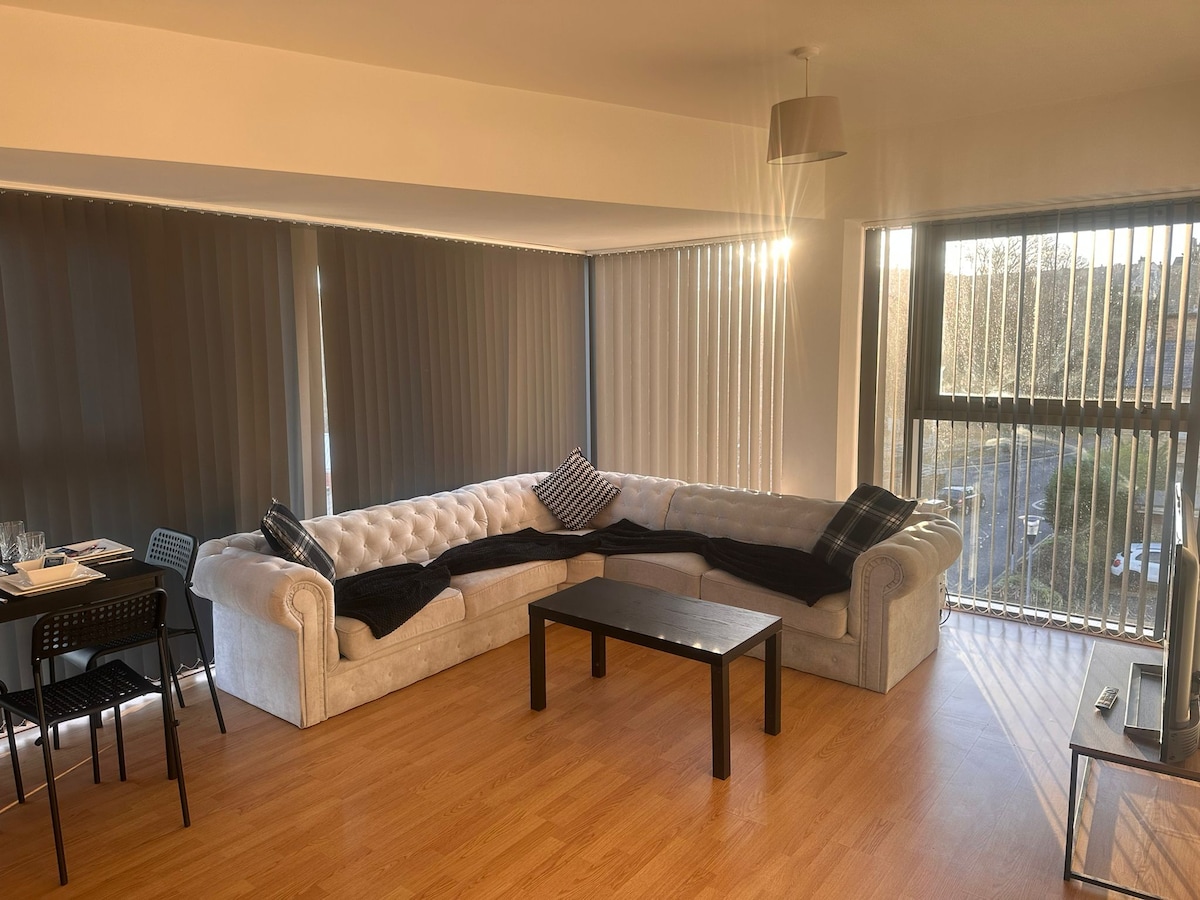 A cozy living area features a large, L-shaped sectional sofa, complemented by a low coffee table in front. Natural light filters through expansive windows with vertical blinds, illuminating the space. A dining table with black chairs is visible nearby, enhancing the room's functionality.