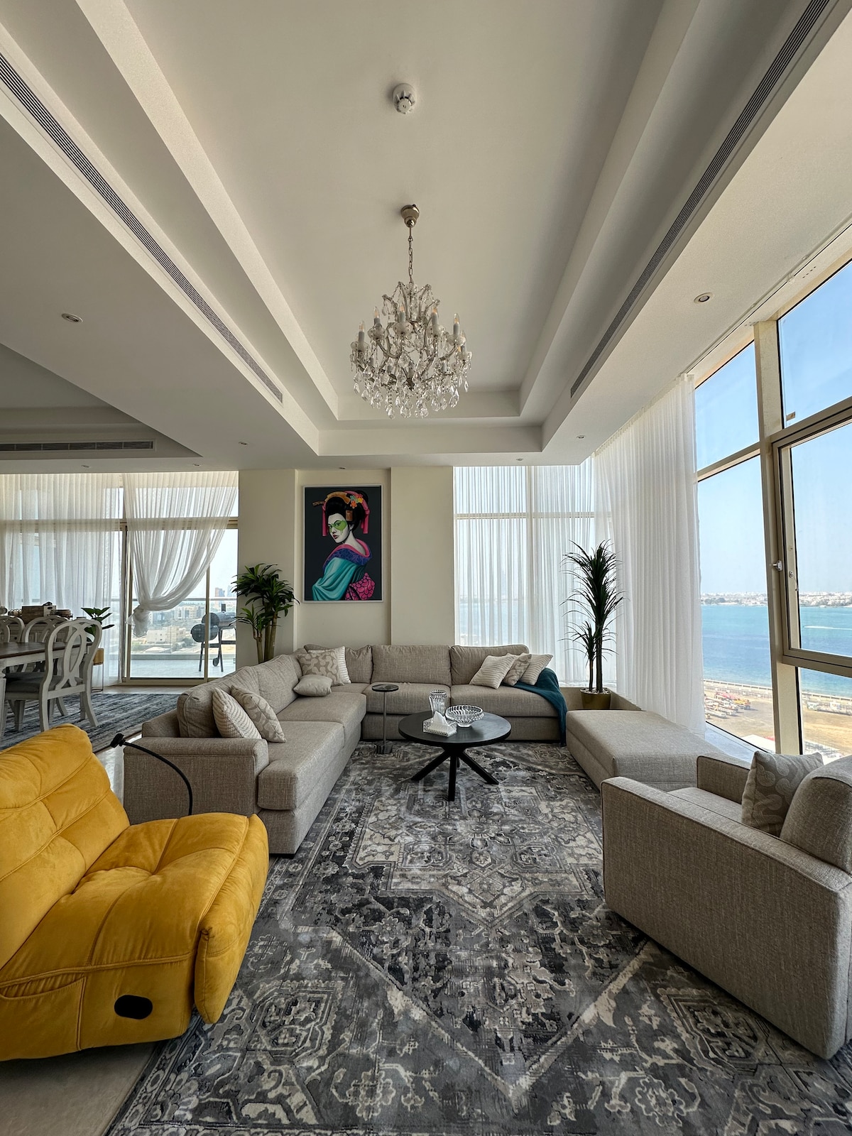 A spacious living area features large windows offering views of the sea. A plush sectional sofa and a vibrant yellow chair are positioned around a central coffee table, complemented by an area rug with intricate patterns. Decorative plants add a touch of greenery to the space.