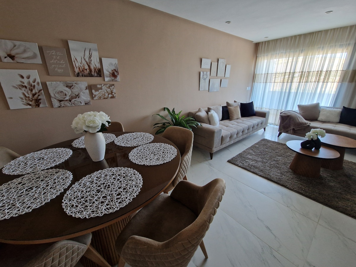 A dining area features a round table surrounded by plush chairs, with decorative placemats arranged neatly on the table. A comfortable seating arrangement includes two sofas, positioned against a wall adorned with artwork. Natural light enters through large windows, creating a bright atmosphere.