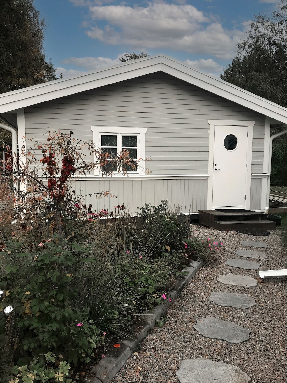 A charming guesthouse is depicted, featuring a light grey exterior with white accents. A round window is visible beside the entrance door, which is framed by lush greenery and colorful flowers lining the pathway. Stone stepping stones lead to the door, enhancing the inviting aspect of the entrance.