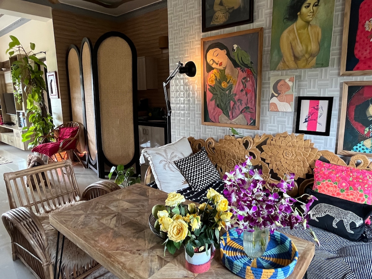 A cozy living space features a wooden dining table surrounded by intricately designed seating. Colorful artwork adorns the walls, while a vibrant flower arrangement enhances the ambiance. Natural light filters through the space, complemented by greenery and decorative elements, creating a warm and inviting setting.