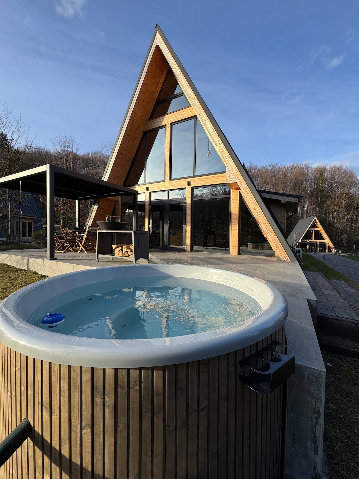 An A-frame structure is shown with large glass windows reflecting natural surroundings. A circular hot tub with clear water is positioned in the foreground on an outdoor patio. A shaded seating area and additional buildings are visible in the background against a clear sky.