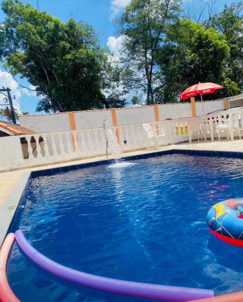 The pool area features a clear blue swimming pool surrounded by a tiled deck. A colorful inflatable ring floats on the water. A bright red umbrella stands nearby, providing shade, while tall trees create a backdrop of greenery.