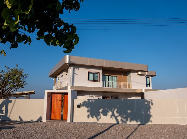 A modern two-story house is displayed against a clear blue sky. The structure features a combination of large windows and a spacious balcony, while a wooden door invites entry. A neatly landscaped area is visible in front, enclosed by a low wall.