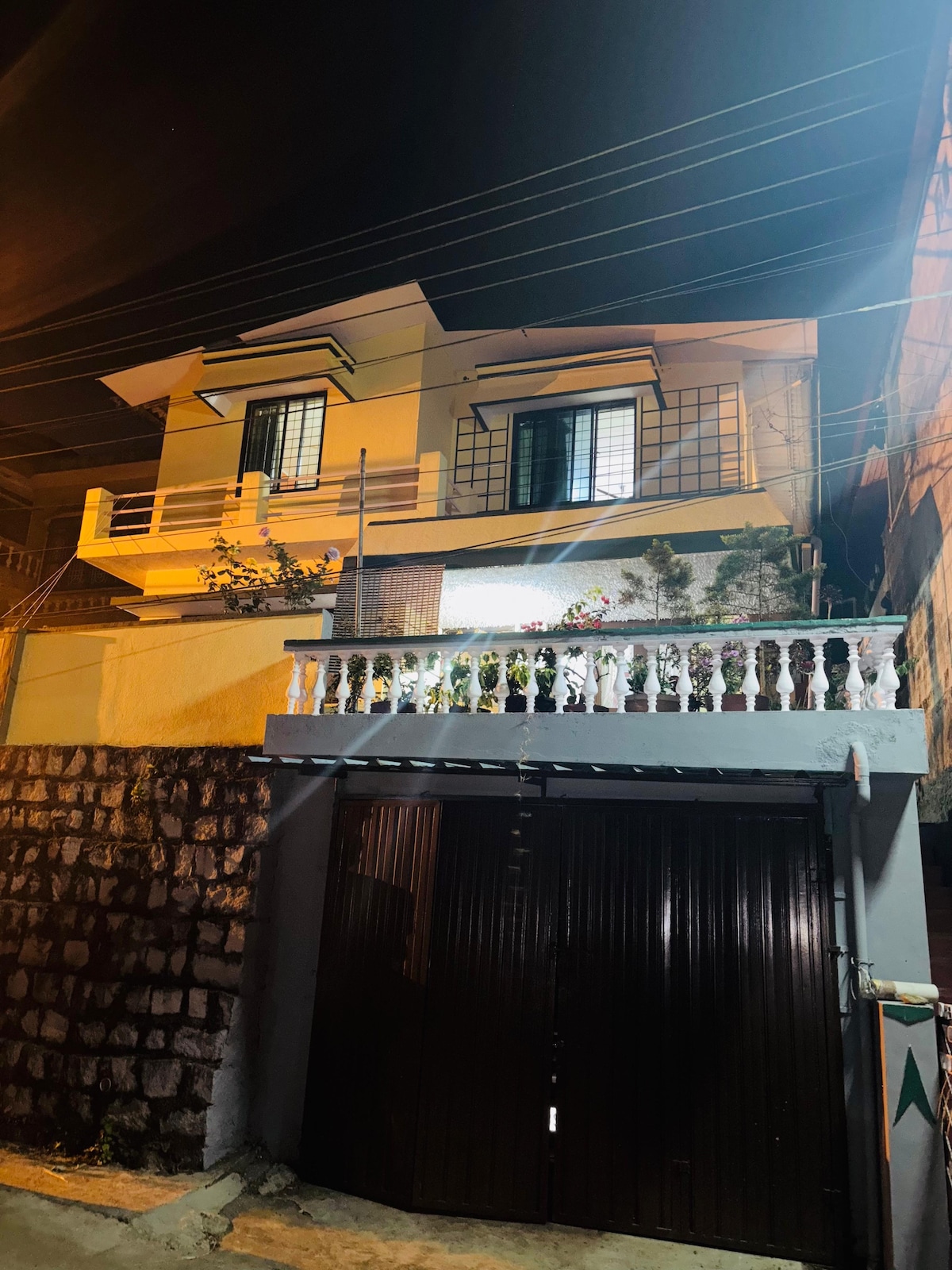 A well-lit two-story building stands at night, featuring a balcony with decorative railings and potted plants. The entrance is highlighted by warm lighting, emphasizing the inviting façade. The dark garage door contrasts with the bright home above.