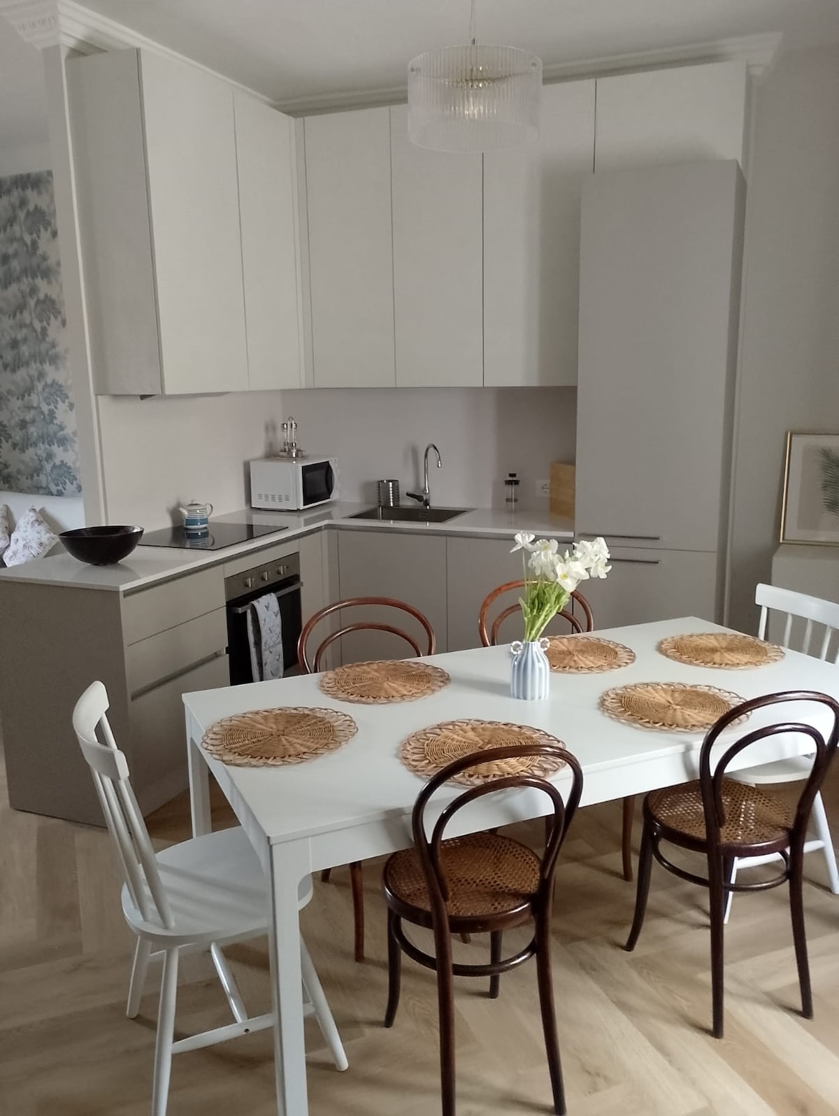 A modern kitchen area is depicted, featuring a light-colored table set for six with woven placemats. A minimalist cabinetry design offers ample storage space. Kitchen appliances, including a microwave and oven, are visible, alongside a small bowl and a vase with flowers on the table.