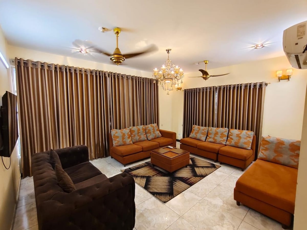 The living room features a brown and orange color palette with a large, cozy sofa set and a matching ottoman. A stylish chandelier hangs from the ceiling, and multiple ceiling fans are present. Elegant curtains frame the windows, complementing the tiled floor.