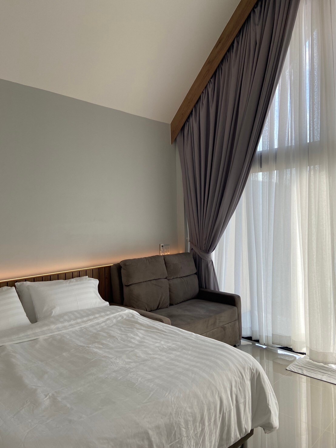A bright bedroom features a spacious bed with white linens, complemented by a comfortable adjustable sofa in the corner. Large windows allow natural light to flow in, with sheer curtains softly diffusing the sunlight.