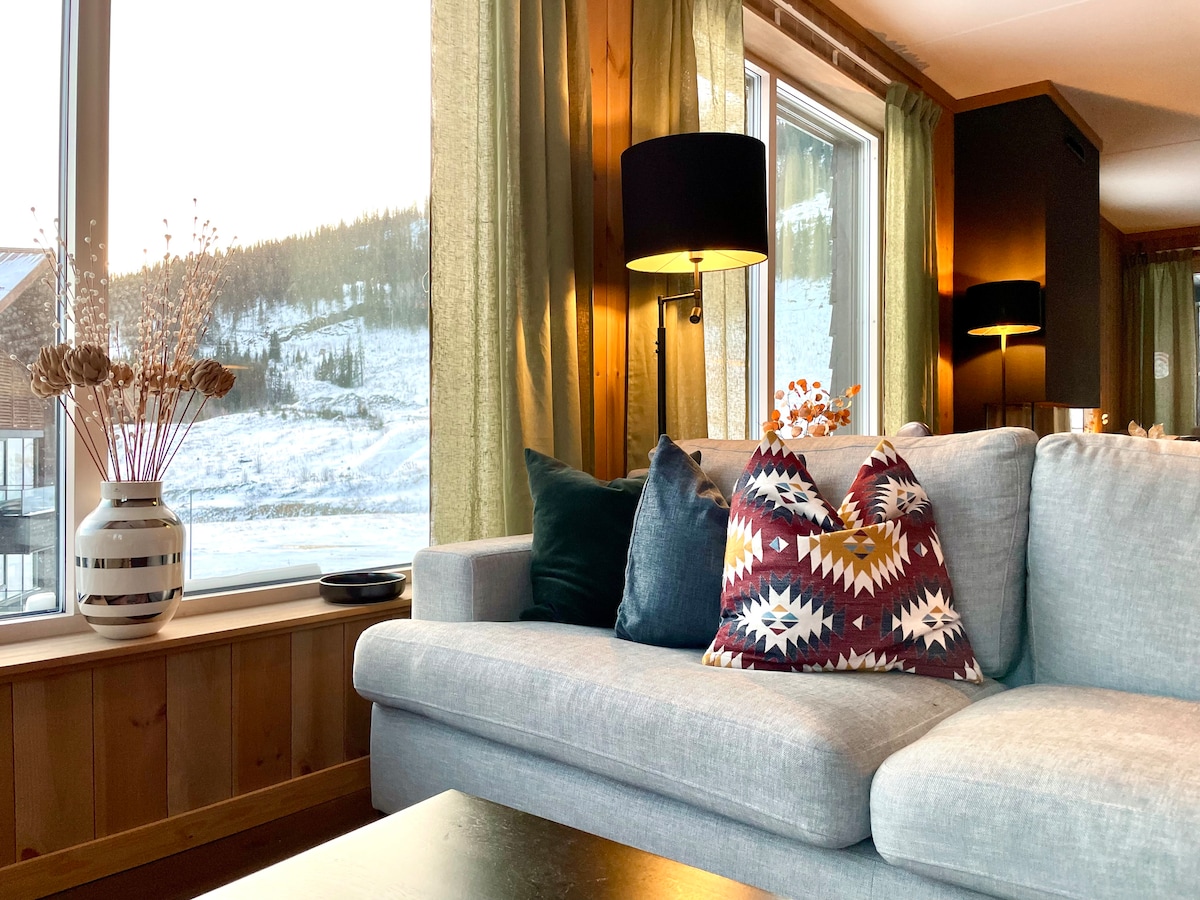 A cozy living area is presented, featuring a soft gray sofa adorned with a decorative pillow. Large windows provide natural light and showcase a snowy landscape. Subtle green curtains frame the windows, complementing the warm lighting from table lamps.