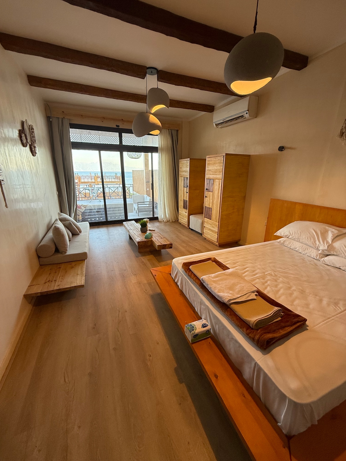 A spacious bedroom features a wooden bed frame with crisp bedding. An inviting seating area with cushions is positioned near a glass door leading to a view. Natural light fills the room, highlighting the wooden furniture and the warm tones of the decor.