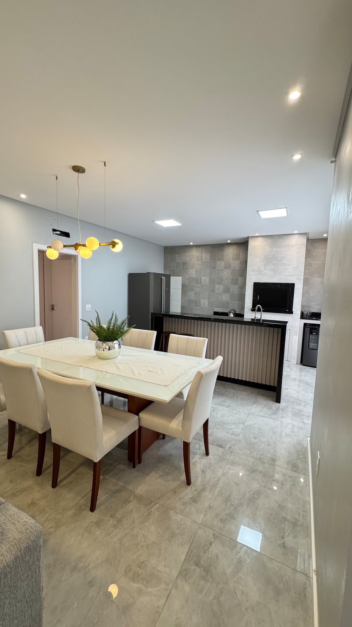 A modern kitchen and dining area feature a light-colored table surrounded by cream chairs. A sleek refrigerator and stove are visible, along with a textured gray backsplash. Overhead lighting creates a bright ambiance, enhancing the space's clean lines and contemporary design.