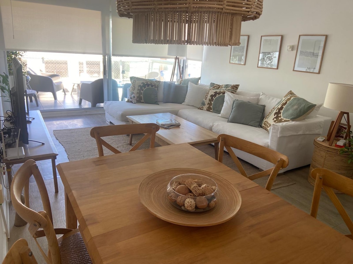 A bright living space is highlighted by a large sectional sofa adorned with various decorative cushions. A wooden dining table is set with a decorative bowl. Natural light fills the room, creating a warm ambiance that is complemented by indoor plants and subtle artwork on the walls.