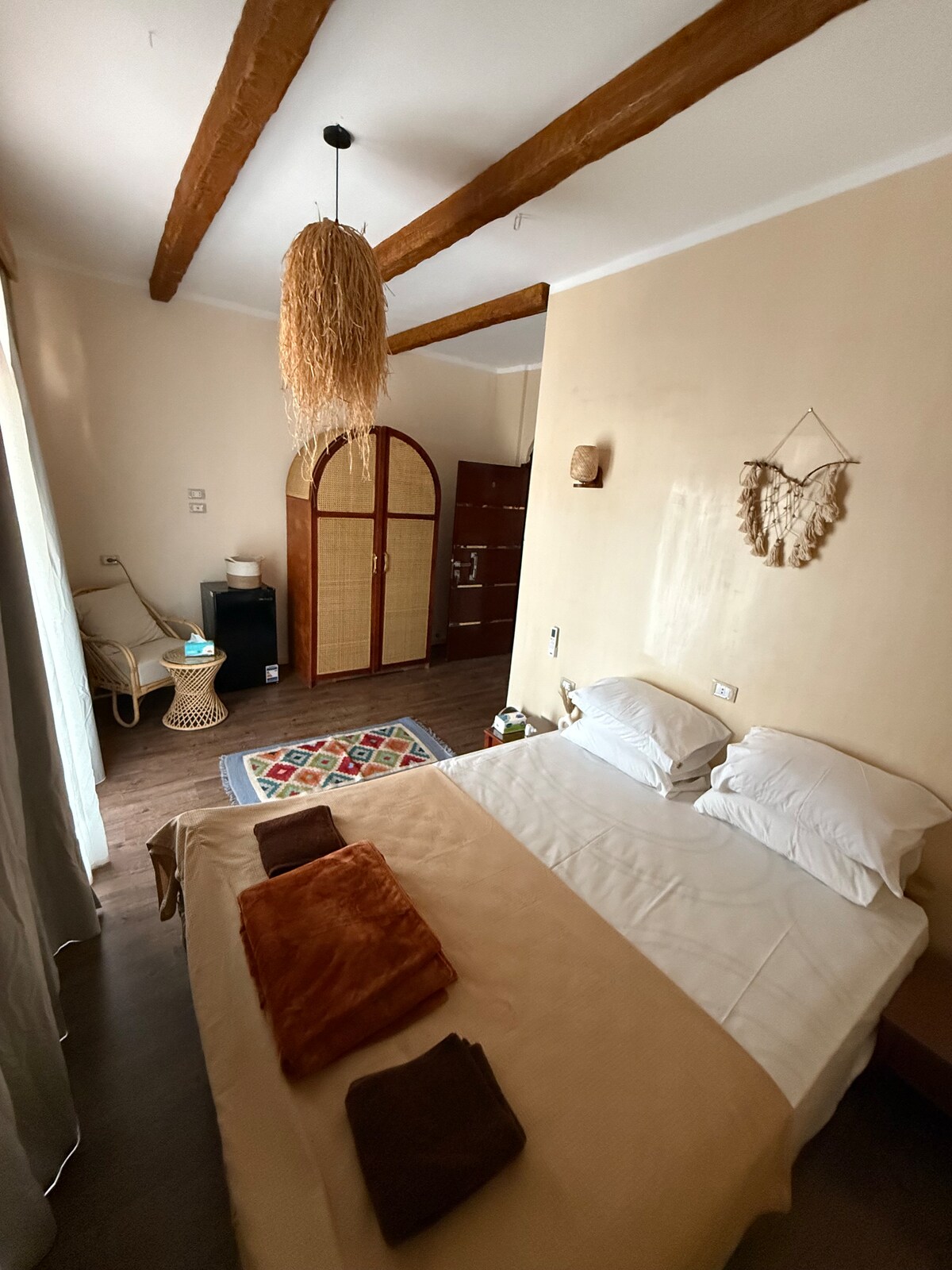 A cozy bedroom features warm wooden beams and soft lighting. A spacious bed is paired with neatly arranged towels. A patterned rug adds color to the floor, while a wooden wardrobe and a small accent chair provide functional decor elements.