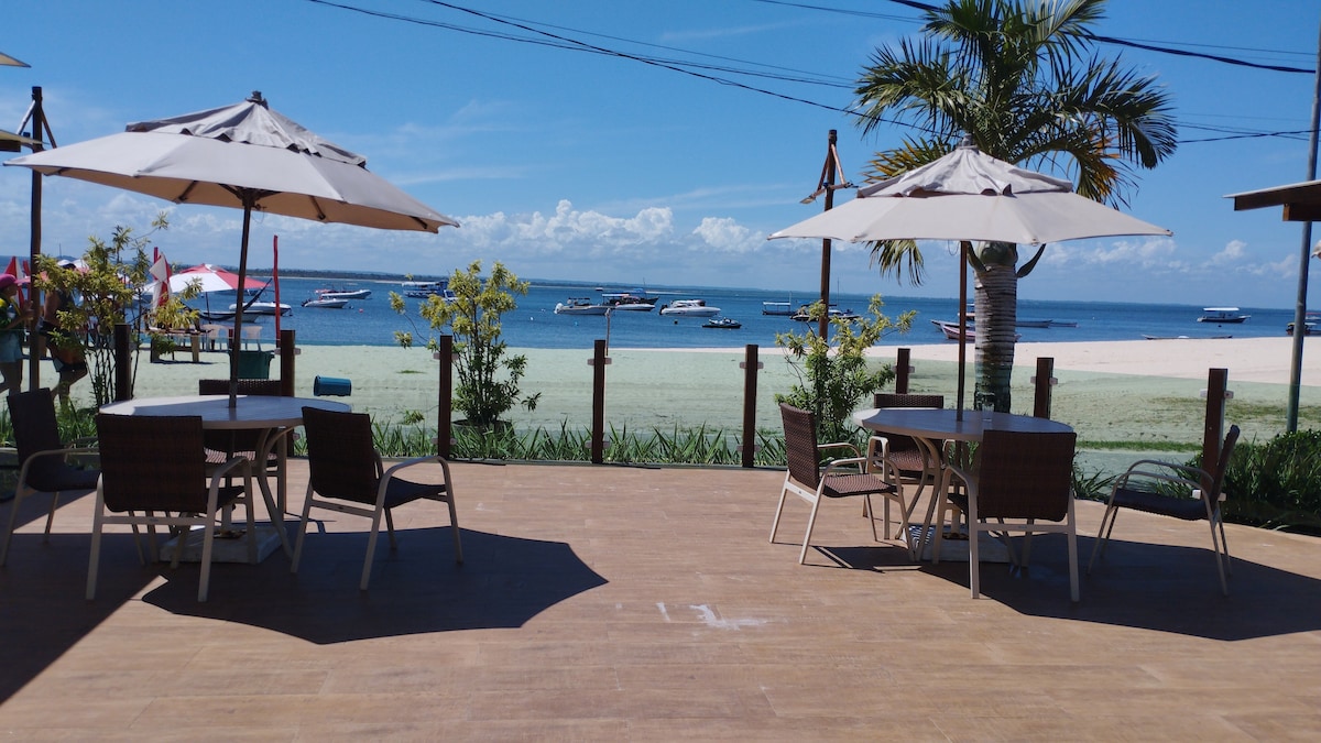 An outdoor patio area is visible, featuring multiple tables and chairs under umbrellas. The background showcases a sandy beachfront and calm waters, with boats anchored nearby. Lush greenery frames the scene, contributing to a relaxed coastal atmosphere.