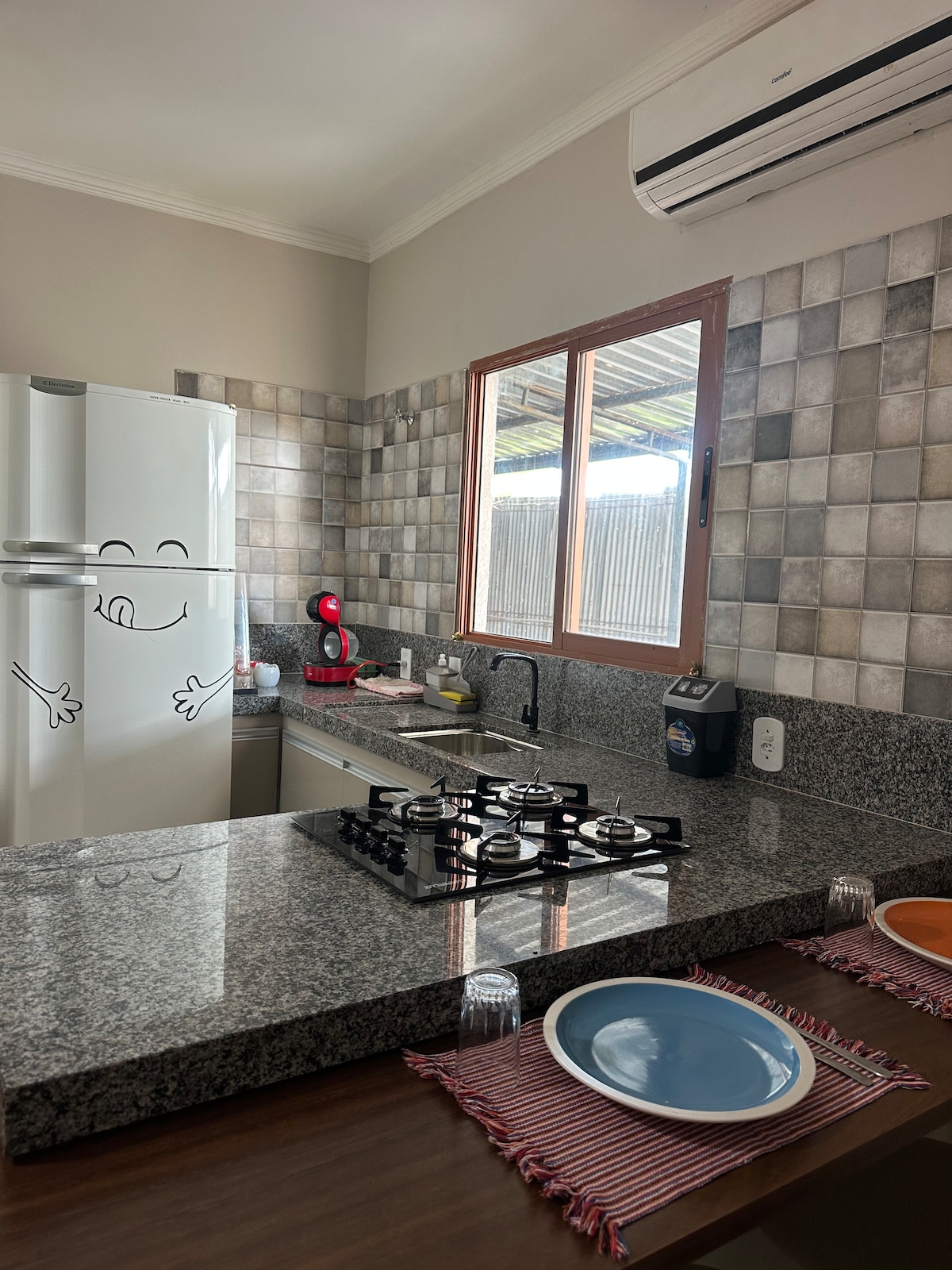 A modern kitchen features a sleek granite countertop and a four-burner gas stove. Natural light enters through a window beside the stainless-steel refrigerator. The space is equipped with utensils and colored plates set on the dining table, adding a practical touch.