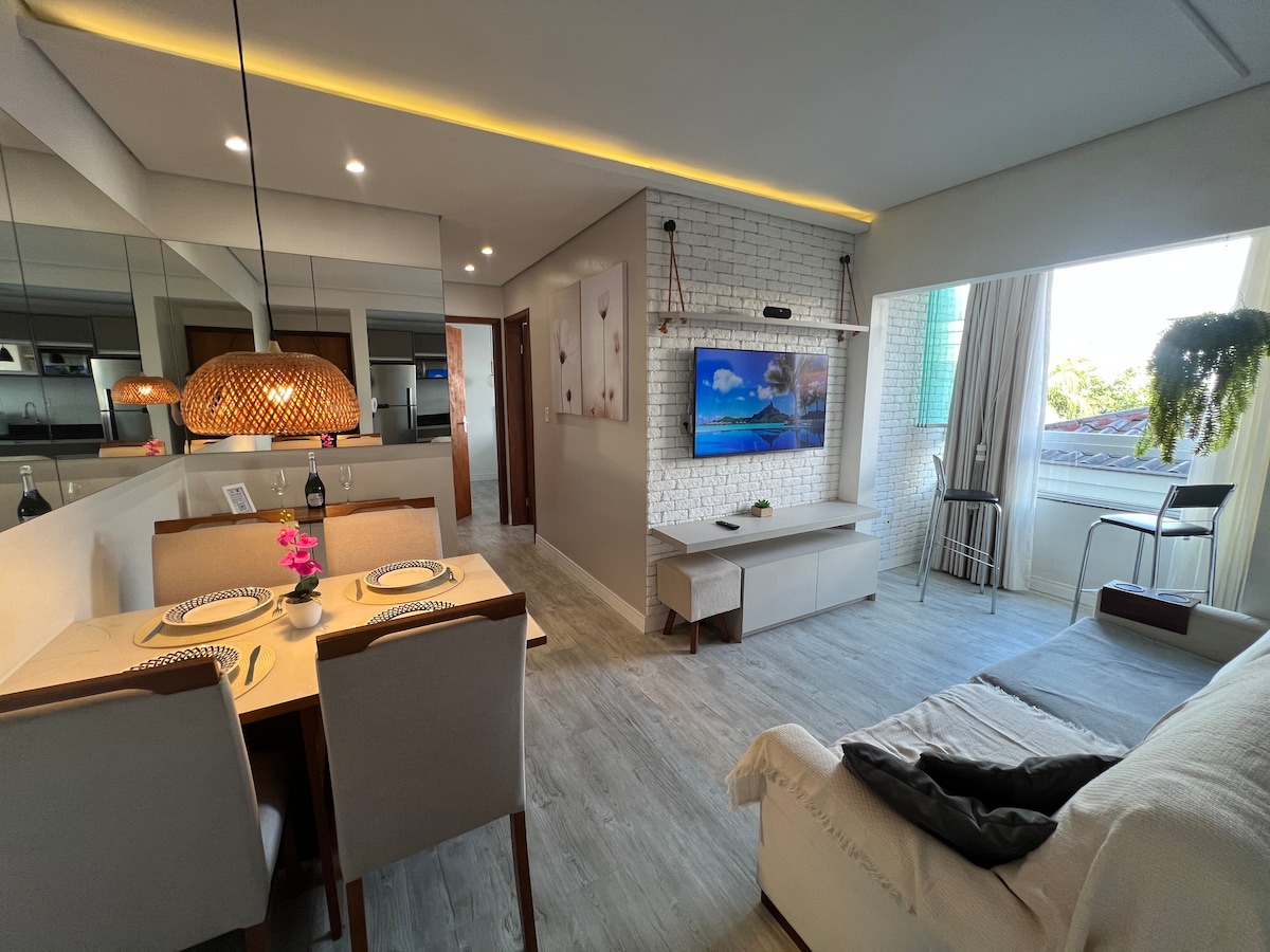 The living and dining area showcases an open layout with a wooden dining table and light-colored chairs. A wall-mounted television is featured prominently, with a stylish decor element above. Natural light enters through large windows, highlighting a comfortable sofa adorned with a soft throw.