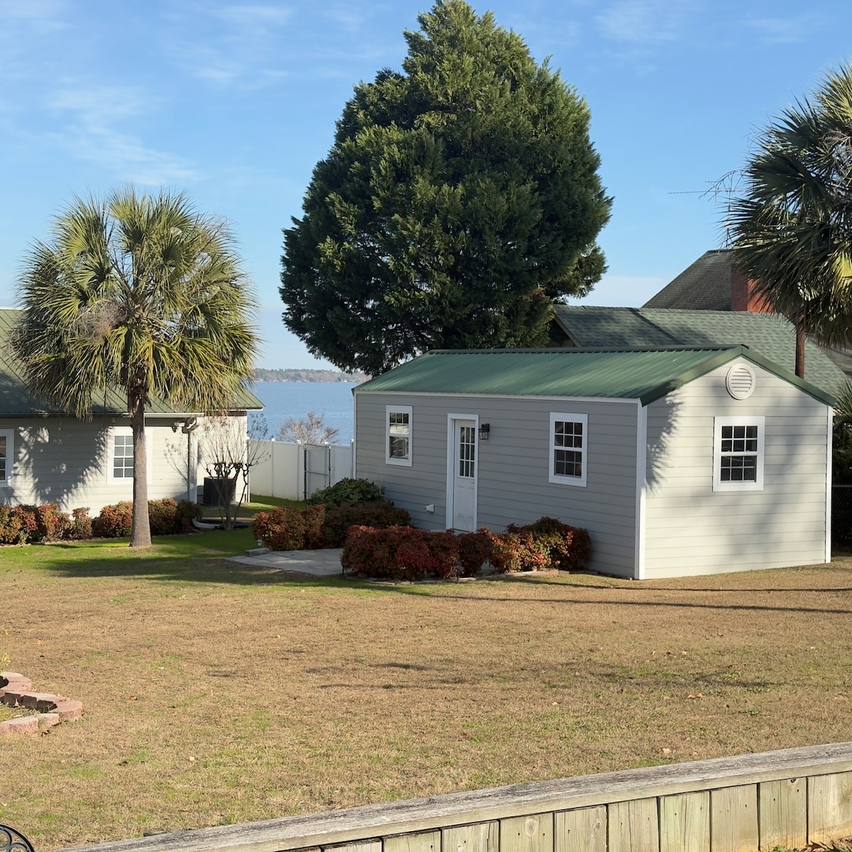 A charming lakefront guest house is set within a well-maintained grassy area, framed by palm trees and vibrant shrubs. The house features a green roof and multiple windows, allowing for ample natural light. The serene lake is visible in the background, enhancing the peaceful environment.