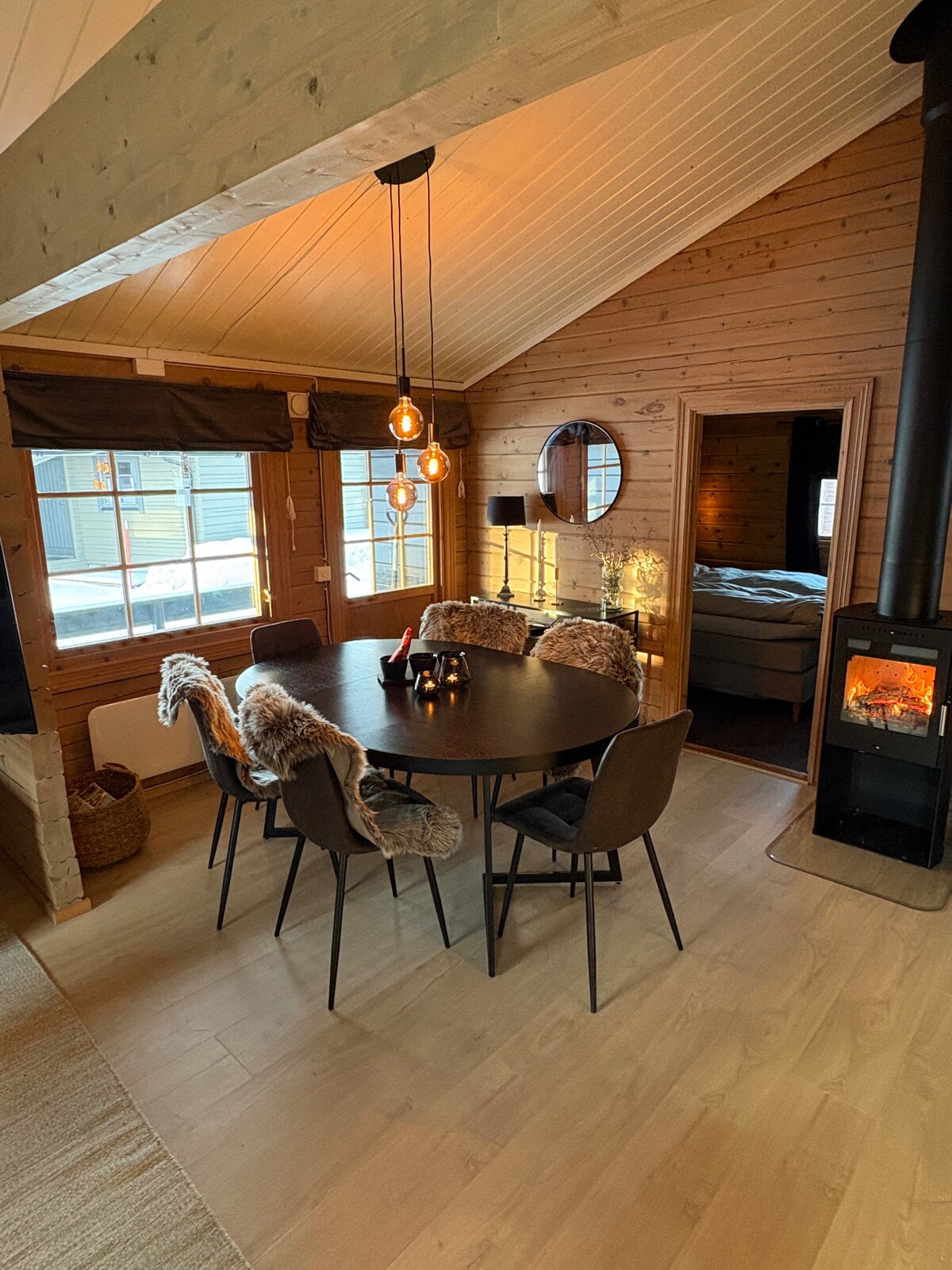 A dining area features a round black table surrounded by six upholstered chairs, enhanced by soft throw blankets. Warm light fixtures hang from a sloped ceiling, illuminating the cozy ambiance. A wood-burning stove stands nearby, adding to the inviting atmosphere of the space.