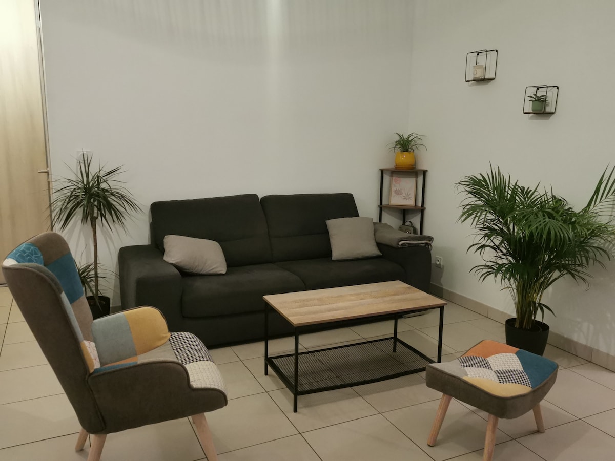 A modern living area features a dark sofa with decorative pillows, positioned across from a wooden coffee table. Two accent chairs with multicolored upholstery add visual interest. Green plants provide a touch of nature, while wall-mounted planters add height and texture to the space.