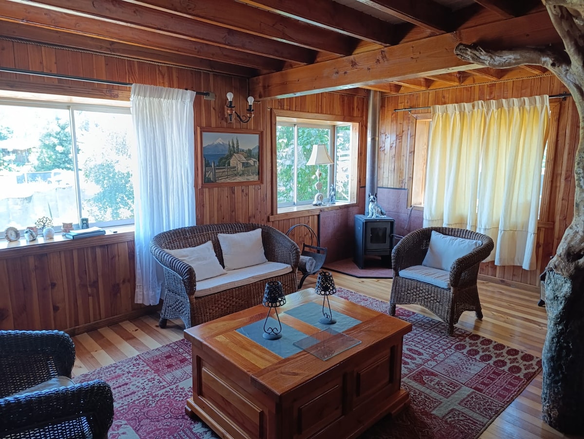 A cozy living area features wooden walls and beams, with natural light streaming through large windows. Two wicker sofas are positioned around a wooden coffee table, with a decorative rug on the floor. A small wood stove adds warmth, complemented by soft curtains.