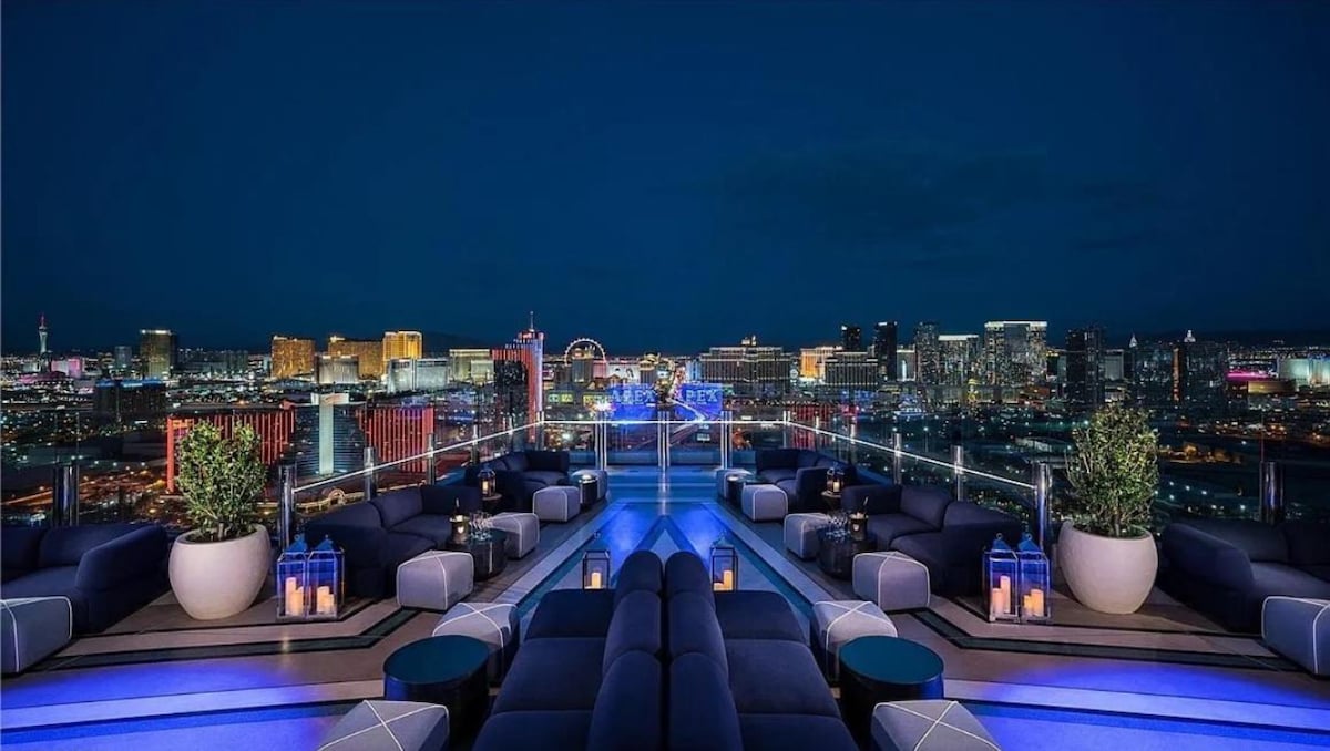A modern rooftop lounge is presented, showcasing low seating arrangements surrounded by potted greenery and lanterns. The vibrant Las Vegas skyline is illuminated in the background, highlighting a blend of colorful lights against the night sky.