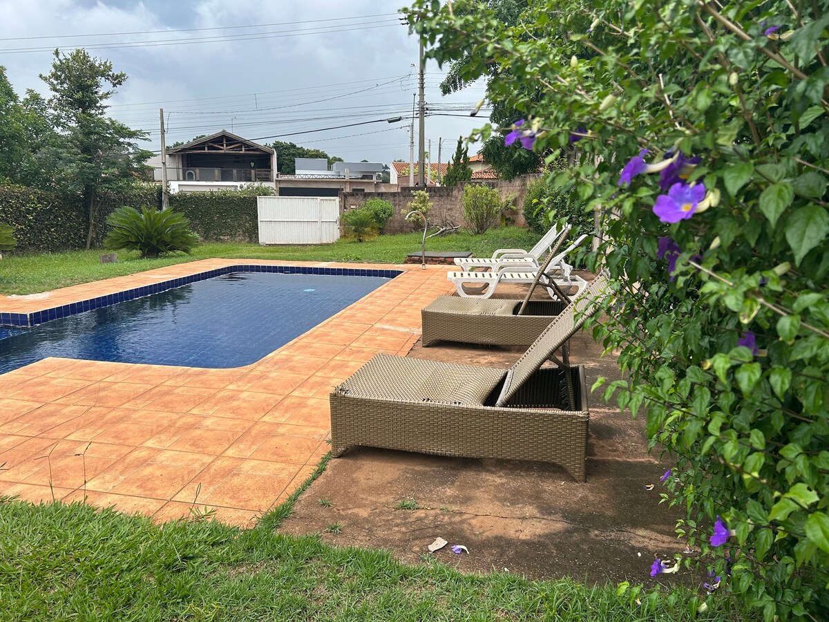 A tranquil pool area features a rectangular pool surrounded by a tiled deck. Several lounge chairs are positioned nearby under the shade of greenery. Vibrant purple flowers add a touch of color, enhancing the serene outdoor setting.