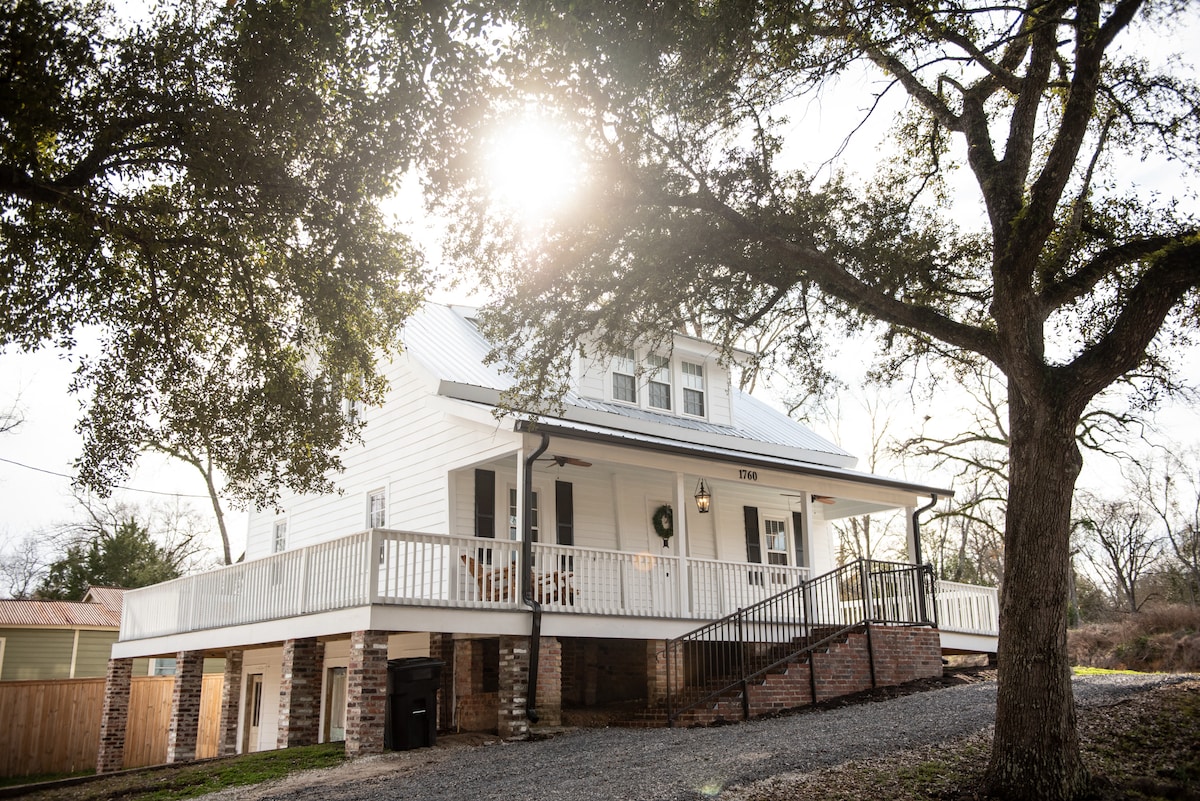 The historic house is viewed from outside, showcasing two stories and a wrap-around porch. Sunlight filters through the trees, illuminating the white exterior. A gravel path leads to the entrance, flanked by greenery, creating an inviting entrance to the home.