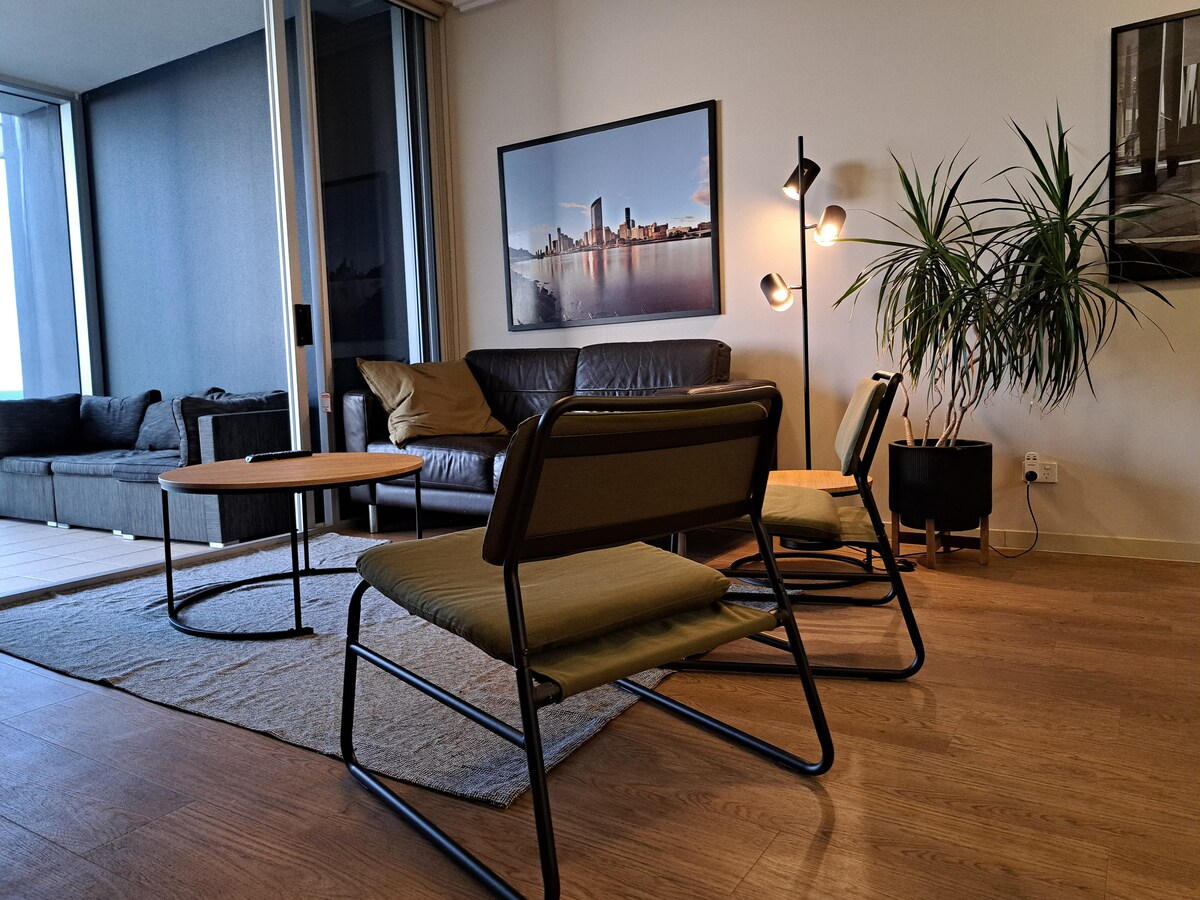A cozy living area displays a modern sofa and two armchairs, arranged around a circular coffee table. Natural light filters through large windows, and a decorative plant adds a touch of greenery. A tasteful cityscape artwork complements the inviting space.