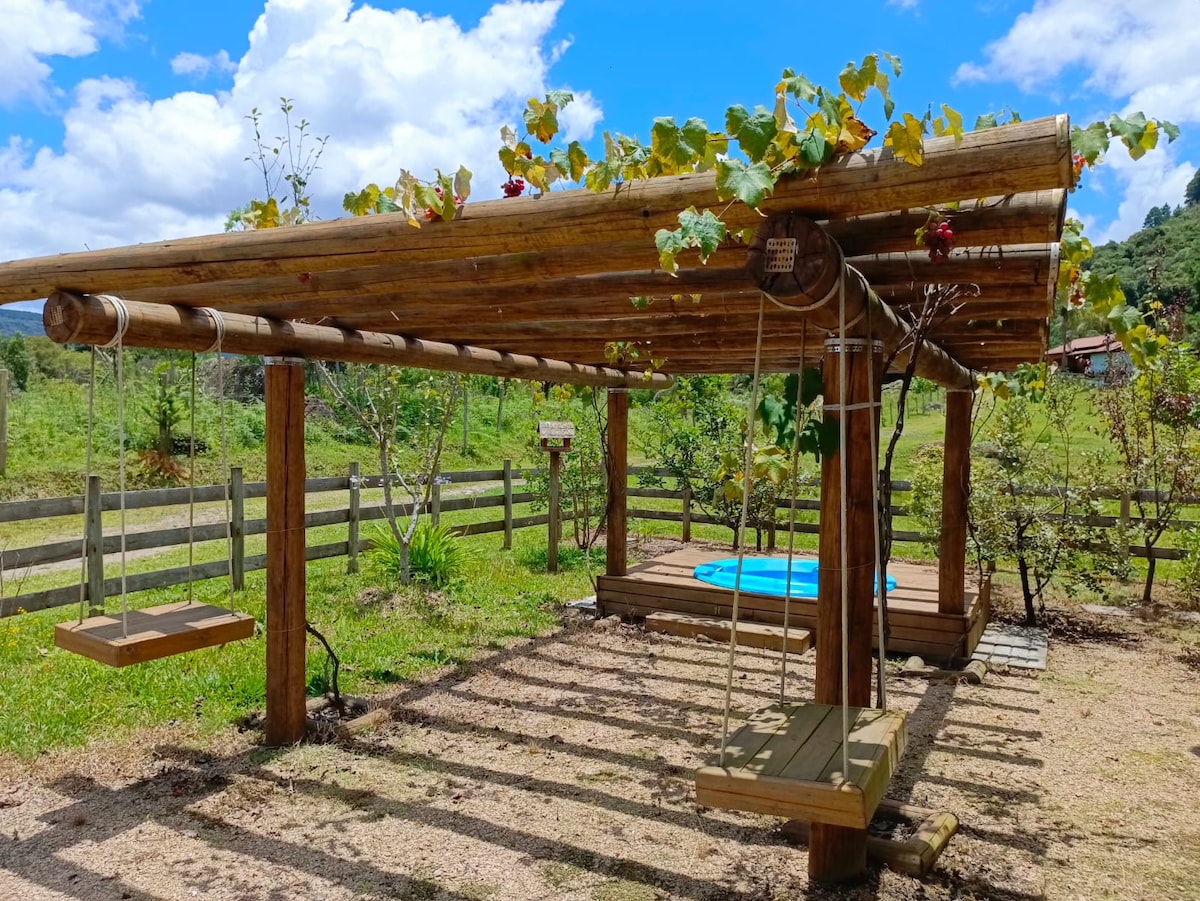A wooden pergola is adorned with climbing vines and provides shade to two swings, positioned on a manicured grassy area. The surrounding landscape features trees and a fenced area, set against a backdrop of blue skies with fluffy clouds.