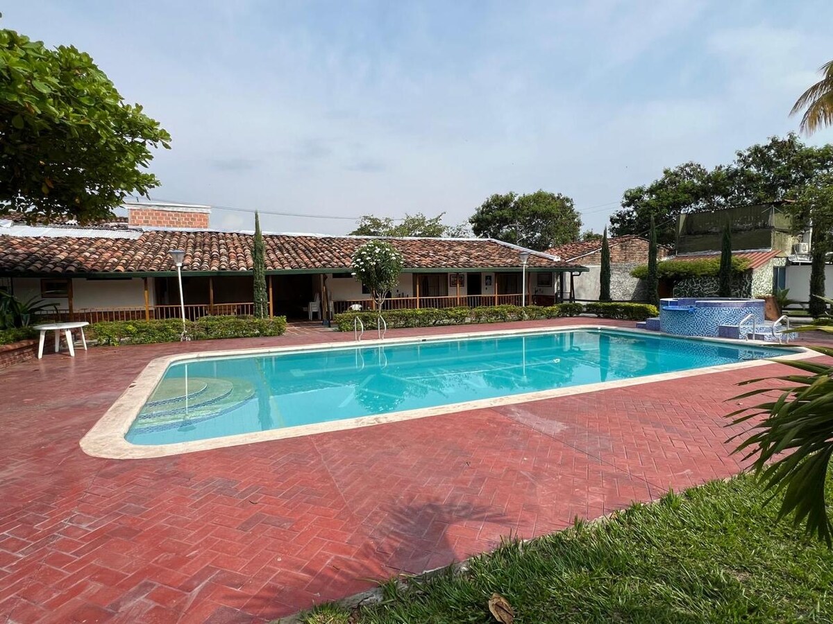 A spacious outdoor pool area features a large swimming pool surrounded by a red-tiled patio. A jacuzzi is situated nearby, accompanied by well-maintained greenery and trees that offer shade. The property exudes a relaxed atmosphere, perfect for family gatherings and warm summer days.