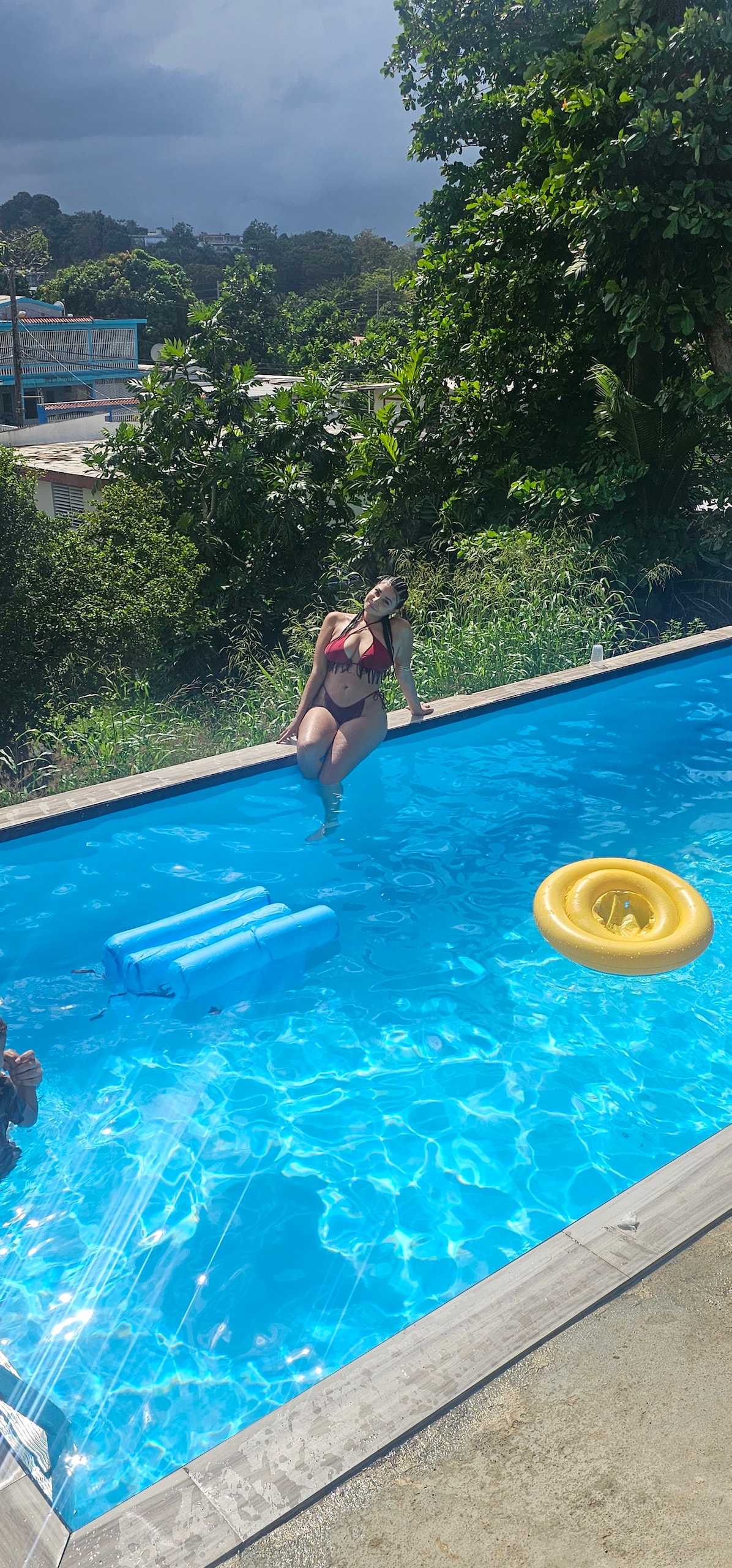 A swimming pool is captured under a clear blue sky, surrounded by lush greenery. A bright floating ring and pool float are visible in the water. Sunlight reflects off the surface, creating a serene and inviting atmosphere for relaxation.