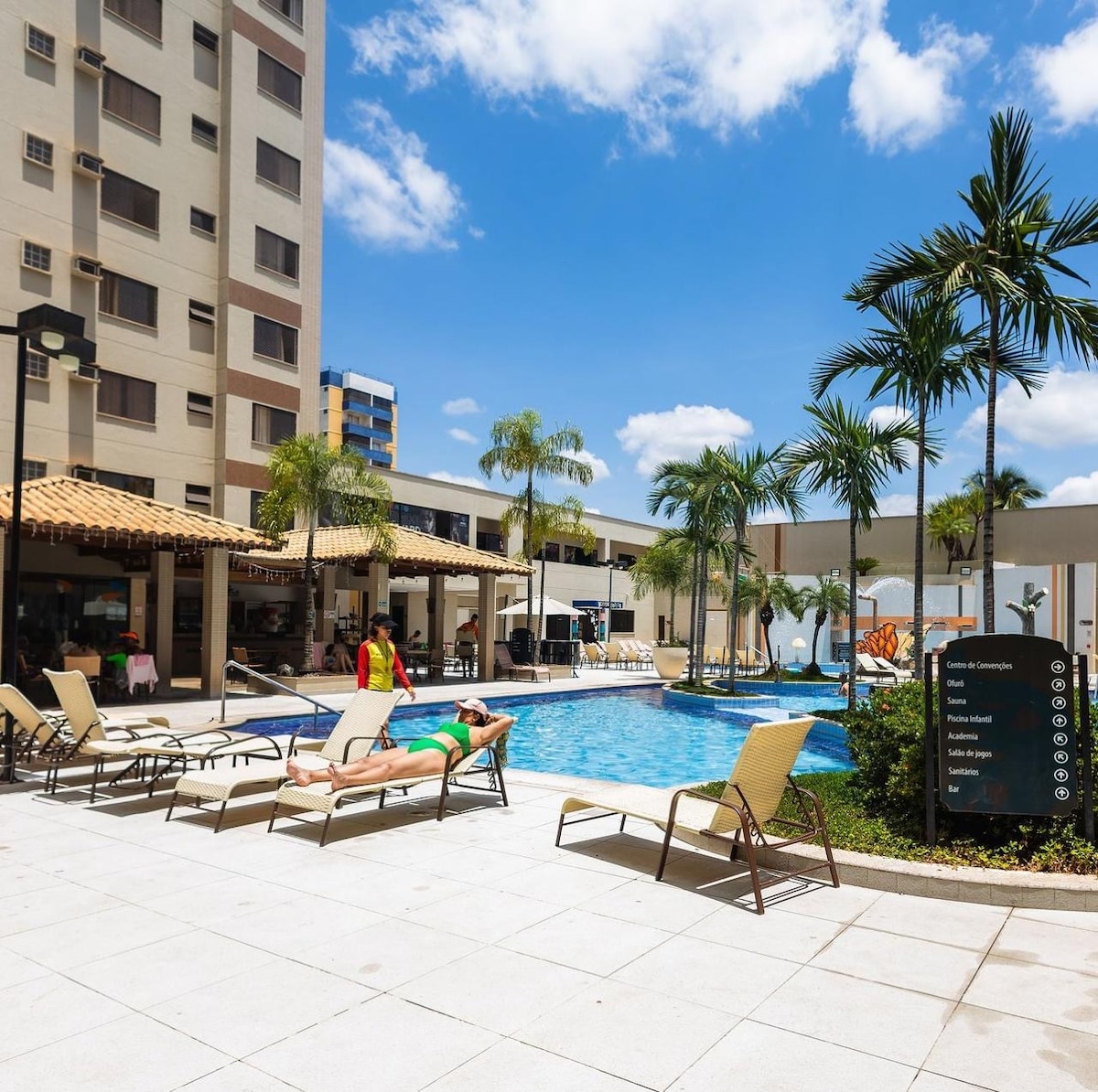 The image shows a sunny outdoor area featuring a swimming pool surrounded by lounge chairs. Tropical plants are visible near the pool, and a covered seating area is located in the background. Clear blue skies and bright sunlight enhance the inviting atmosphere.