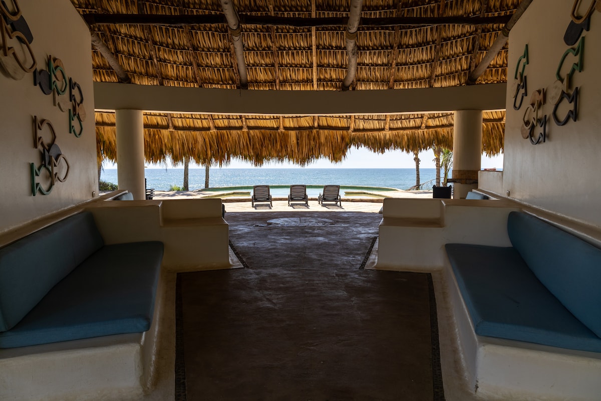 A spacious open area features two built-in benches with blue cushions, providing a relaxed setting. The view reveals a sandy beach and calm ocean waters beyond, framed by a thatched roof. Sun loungers are positioned towards the water for leisure and enjoyment.