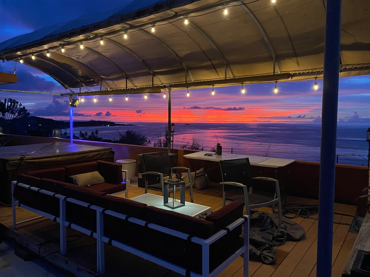 A rooftop terrace features comfortable seating arranged around a center table, with sunset hues illuminating the ocean in the background. Warm string lights provide soft lighting, enhancing the outdoor space's inviting atmosphere as the day transitions into evening.