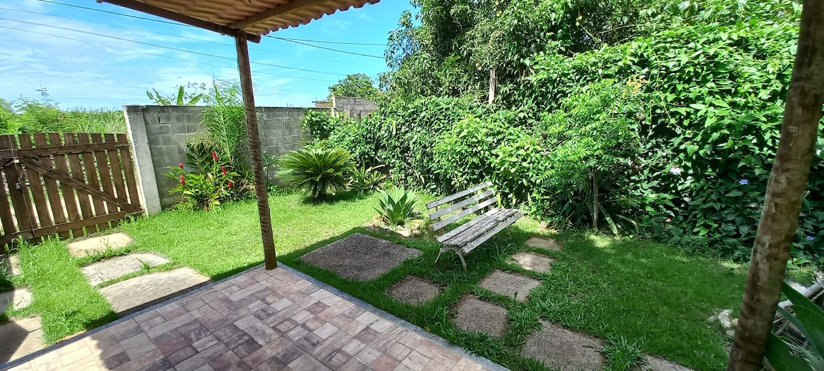 A garden space is showcased, featuring a stone pathway surrounded by lush greenery. A wooden bench provides seating, situated on grass with vibrant plants nearby. The area is shaded by a roof overhang, allowing for a comfortable outdoor experience.