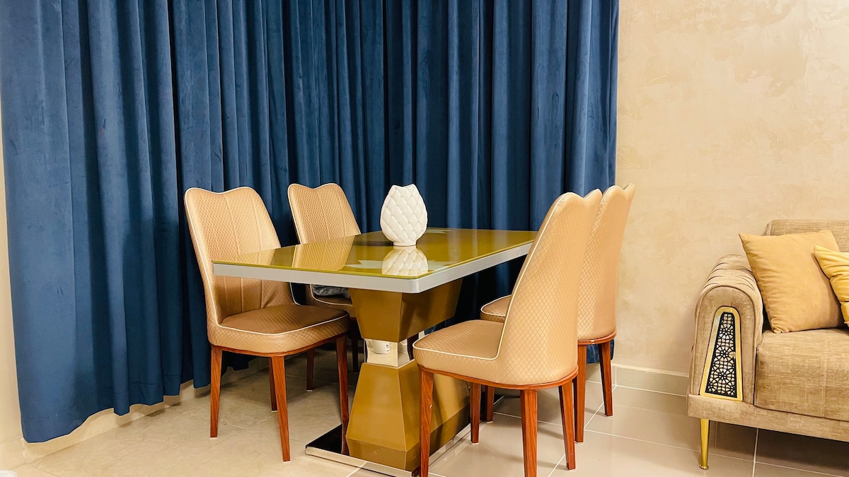 A modern dining area features a glass-top table surrounded by four beige upholstered chairs. A white decorative vase sits centrally on the table. Floor-length dark blue curtains frame the space, enhancing the overall elegance, while a light-colored sofa with decorative cushions is positioned nearby.
