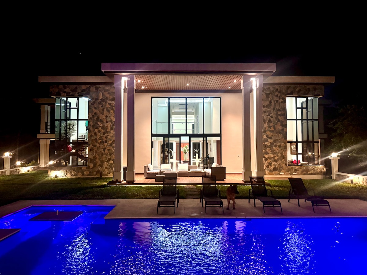 The villa's exterior is illuminated at night, showcasing large glass windows framed by stone accents. A turquoise pool reflects the lights, while lounge chairs are arranged around it. The well-lit patio invites relaxation and enjoyment of the outdoor space.