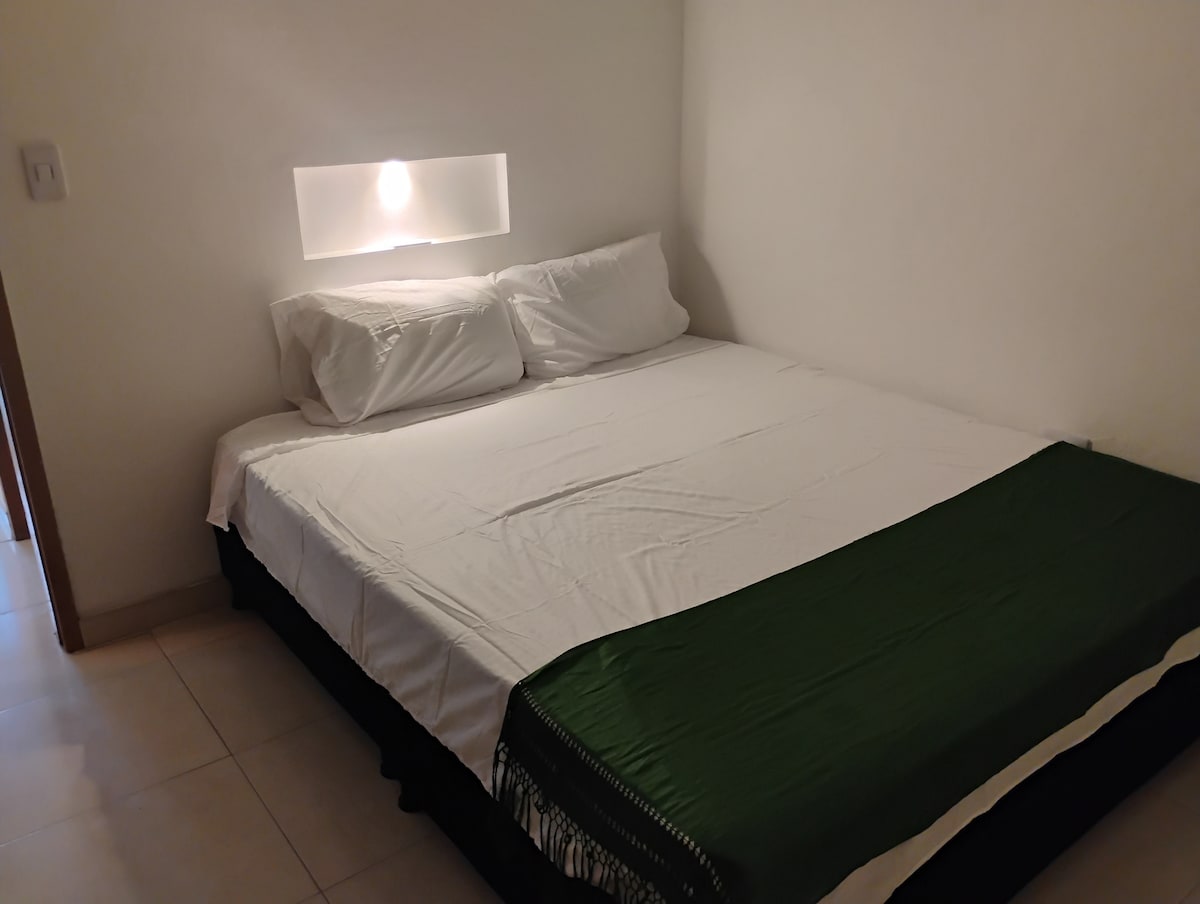 A spacious bed is dressed with crisp white linens and accented by a deep green throw. Soft lighting is provided by a wall-mounted lamp, illuminating the serene environment. The minimalistic design and neutral walls contribute to a calming atmosphere.