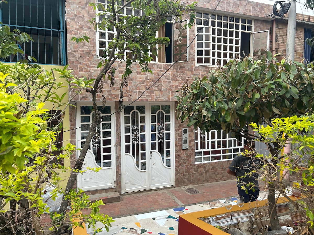 A charming brick façade features two white double doors framed by green accents. Large windows allow natural light to enter the space. Lush foliage surrounds the entrance, complementing the garden's vibrant colors adding to the overall inviting atmosphere of the area.