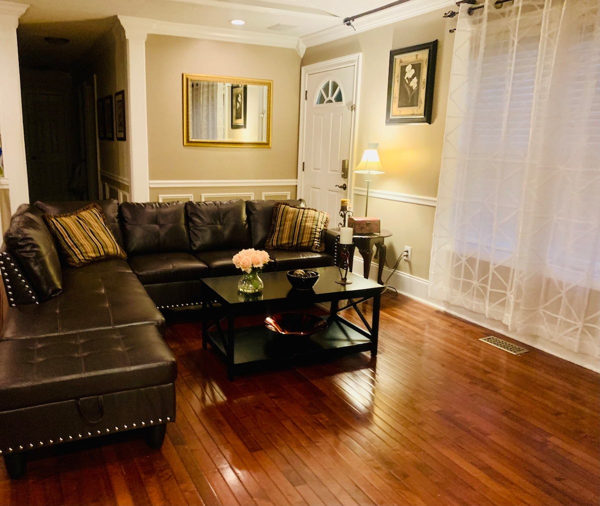 A spacious living area features a dark leather sectional sofa with decorative pillows, facing a glass coffee table placed on polished hardwood flooring. Natural light enters through sheer curtains, illuminating the warm-toned walls adorned with framed artwork.